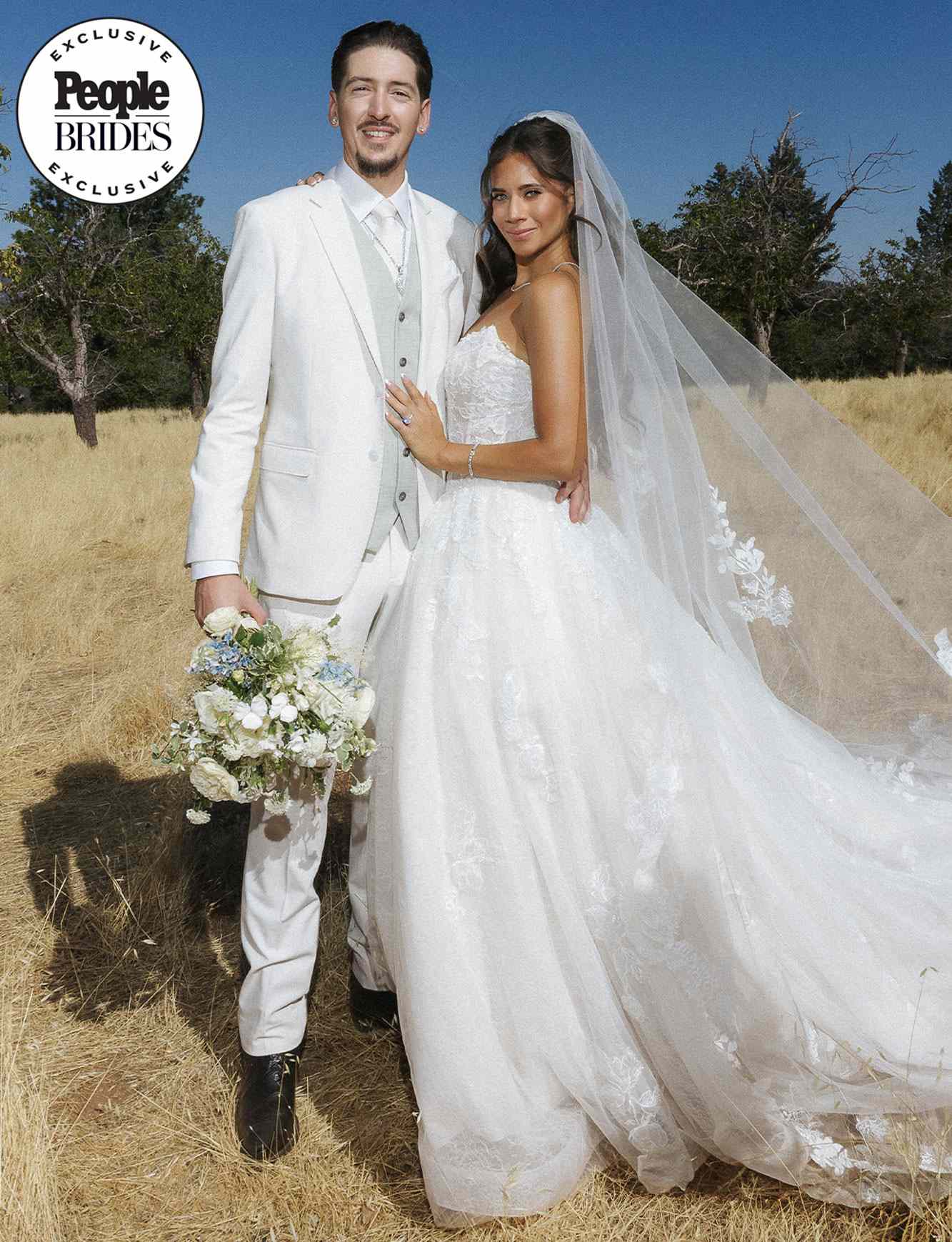 A couple posing outdoors in wedding attire the bride holds a bouquet and wears a veil