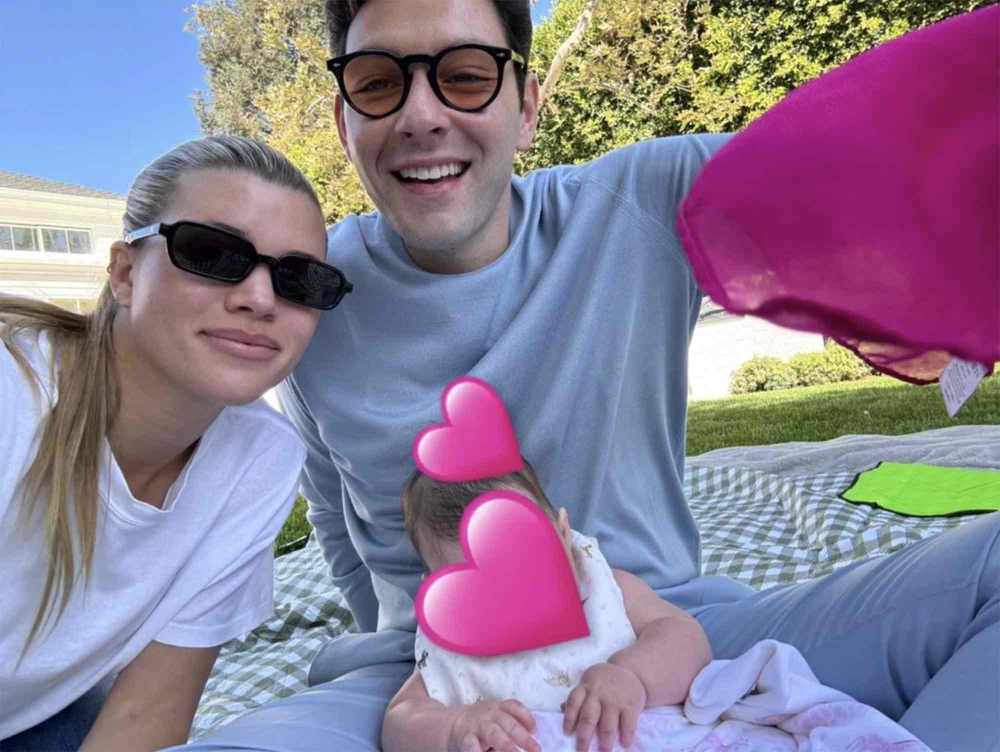 Sofia Richie sitting outdoors with others on a picnic blanket, child concealed by pink heart icons